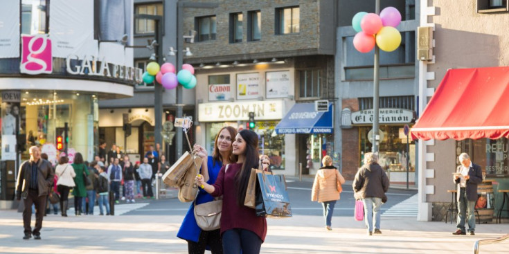 Dues dones amb bosses de compres de diferents comerços del Principat es fan una ‘selfie’ a la plaça de la Rotonda.